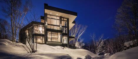A modern house with large windows and a flat roof, surrounded by snow.