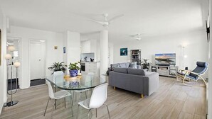 A modern living room with a glass dining table, white chairs, a grey sofa, a TV, and a ceiling fan. - Ocean Waves - boasting beautiful beach views (Nambucca Heads)