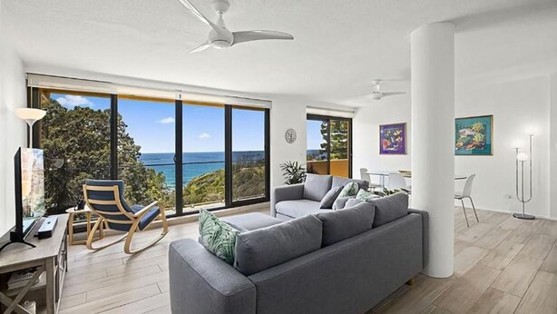 A modern living room with a grey sofa, a desk with a computer, and a view of the ocean through large windows. - Ocean Waves - boasting beautiful beach views (Nambucca Heads)