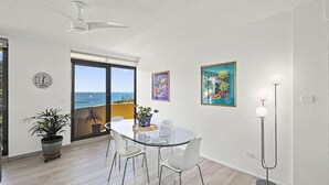 A modern living room with a glass table, white chairs, a potted plant, and artwork on the walls. - Ocean Waves - boasting beautiful beach views (Nambucca Heads)