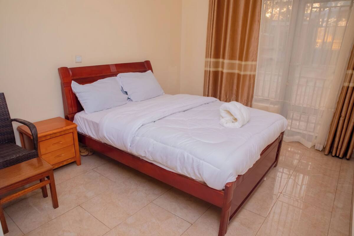 A neatly made bed with white linens and a white towel, a wooden nightstand with drawers, a chair, and a window with curtains.