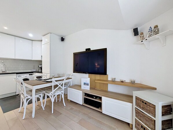 A modern kitchen with white cabinetry, a dining area with white chairs, a flat-screen TV mounted on the wall, and a small shelf with decorative items.