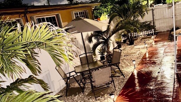 A patio with a table and chairs, surrounded by palm trees and a white fence. - Charming house in terrific Miami with AC, WiFi (North Miami Beach)