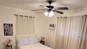A bedroom with a bed, a ceiling fan, a bedside table, a wall-mounted air conditioner, and a window with curtains. - Charming house in terrific Miami with AC, WiFi (North Miami Beach)