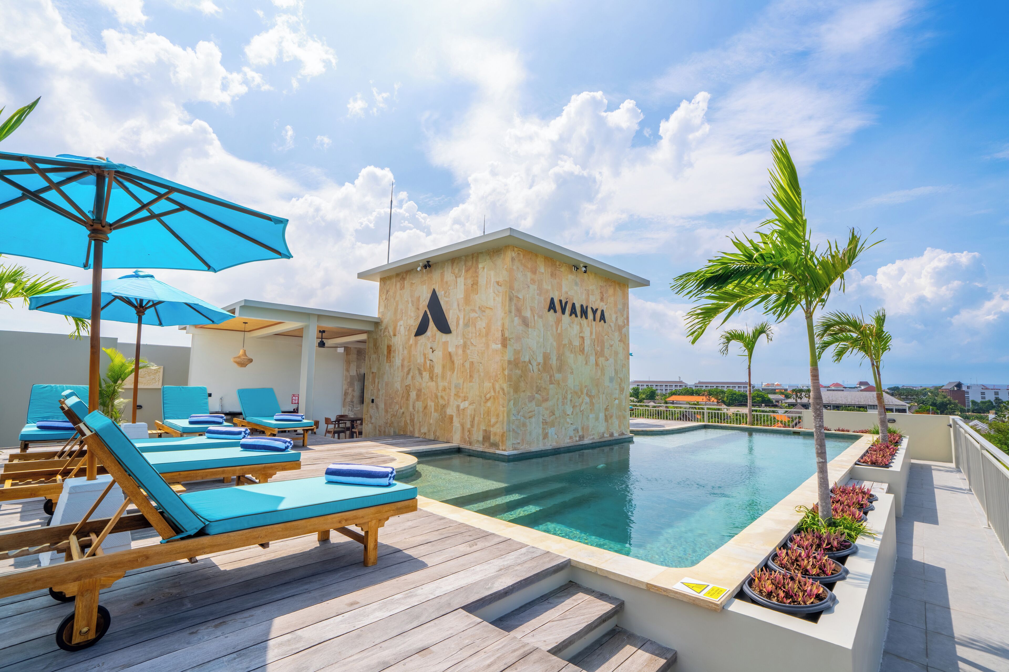 Zona de piscina en la azotea con sillones de descanso, una sombrilla azul y un edificio identificado como "AVANYA".