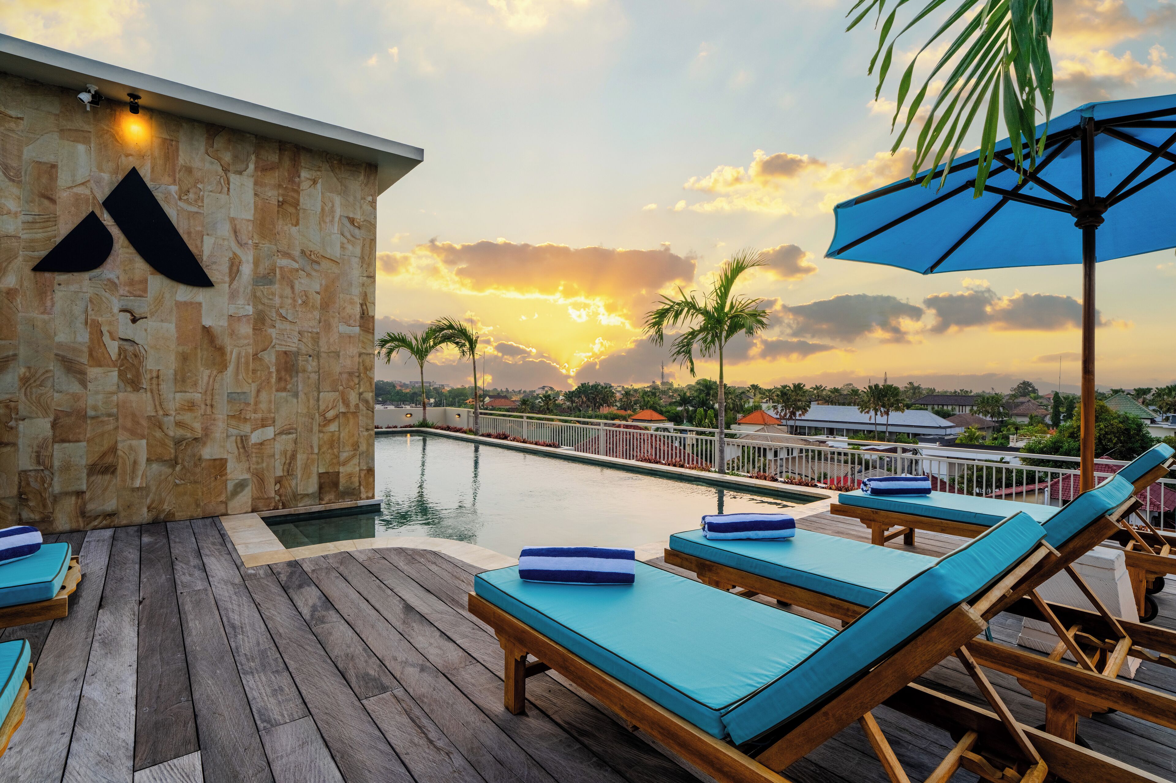 Une terrasse en bois avec des chaises longues et une piscine, d’où l’on peut admirer un coucher de soleil sur des palmiers et un paysage urbain lointain.