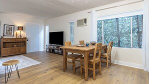 A spacious living room with a wooden dining table, chairs, a TV, and a large window with a view of trees. - Melrose Village Charmer. Walk to world famous shopping and restaurants. (Los Angeles)