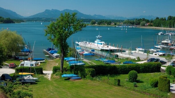 A marina with boats docked, a large white boat in the center, and mountains in the background.