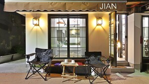 A cozy outdoor seating area with two black chairs and a small table, a storefront with the name "JIAN" illuminated, and a large window with curtains.