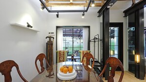 A dining area with a wooden table, chairs, and a fruit platter. There is a large window with curtains, a wooden beam ceiling, and a shelf with a small item.
