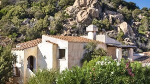Un bâtiment de style méditerranéen traditionnel, avec un toit en tuiles, entouré d’une végétation luxuriante et de collines rocheuses.