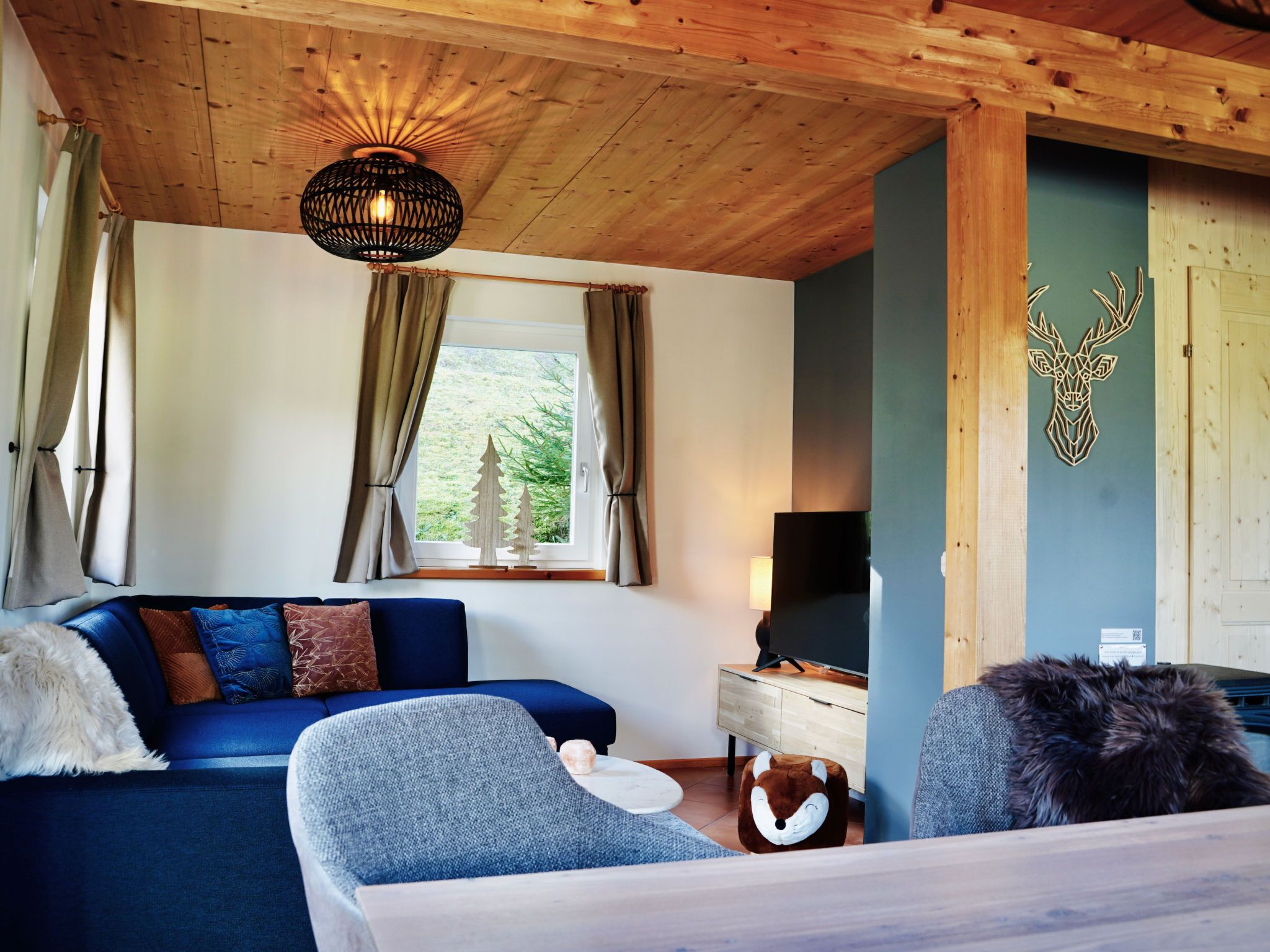A cozy room with a wooden ceiling, a sofa with cushions, a television, and a deer head decoration on the wall.