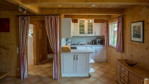 Cocina compacta con armarios blancos, microondas y pileta. Se observa un mueble de madera con un cuenco encima. El ambiente tiene piso de cerámica y paredes de madera.