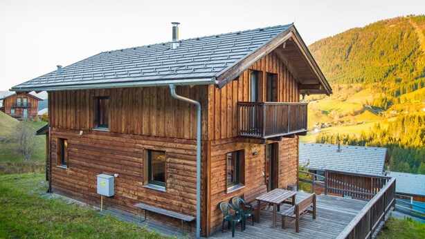 Una cabaña de madera con balcón, terraza con asientos y vistas a un paisaje montañoso.