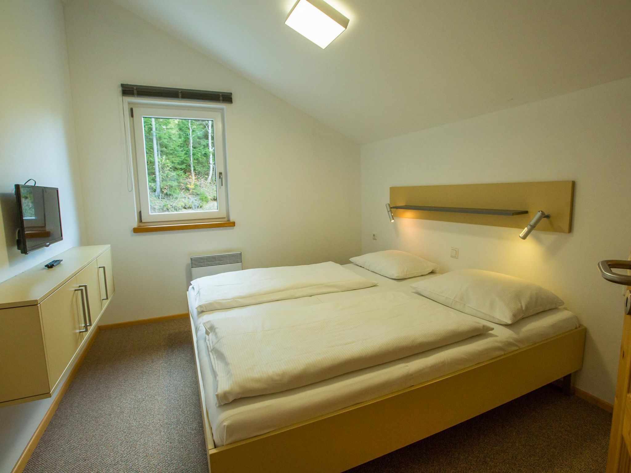 Ein kleines, helles Schlafzimmer mit einem Einzelbett, einem Nachttisch, einem Fernseher und einem Fenster mit Blick auf Bäume.
