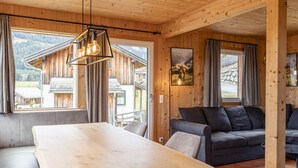 Interior de madera con una mesa de comedor, sofá y ventanas amplias que ofrecen vistas a un paisaje montañoso.