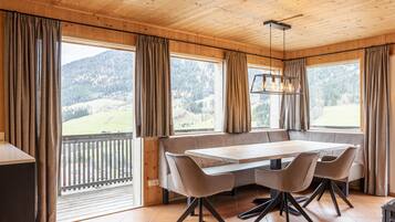 Ein Holzinterieur mit Esstisch, Stühlen und großen Fenstern, die einen Blick auf die Berge ermöglichen.