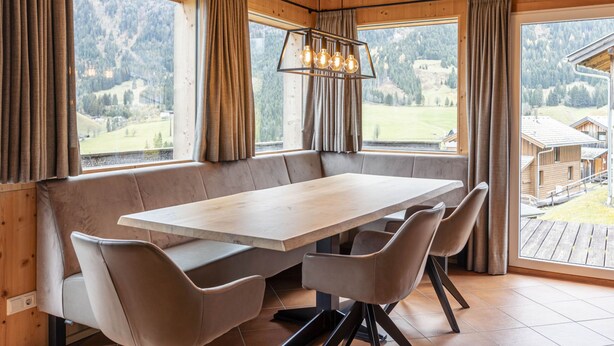 Un comedor con una gran mesa de madera y bancos acolchados, ventanas amplias con cortinas y vistas a un paisaje montañoso.
