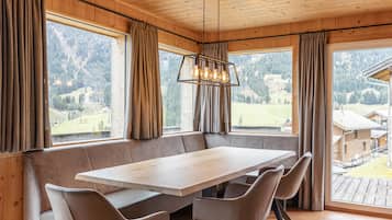 Ein Essbereich mit einem großen Holztisch, gepolsterten Bänken, großen Fenstern mit Vorhängen und Blick auf eine Berglandschaft.