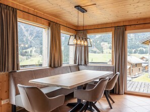 A dining area with a large wooden table and cushioned benches, large windows with curtains, and a view of a mountainous landscape.