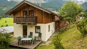 A wooden chalet with a balcony, a deck with chairs, and a mountain backdrop.