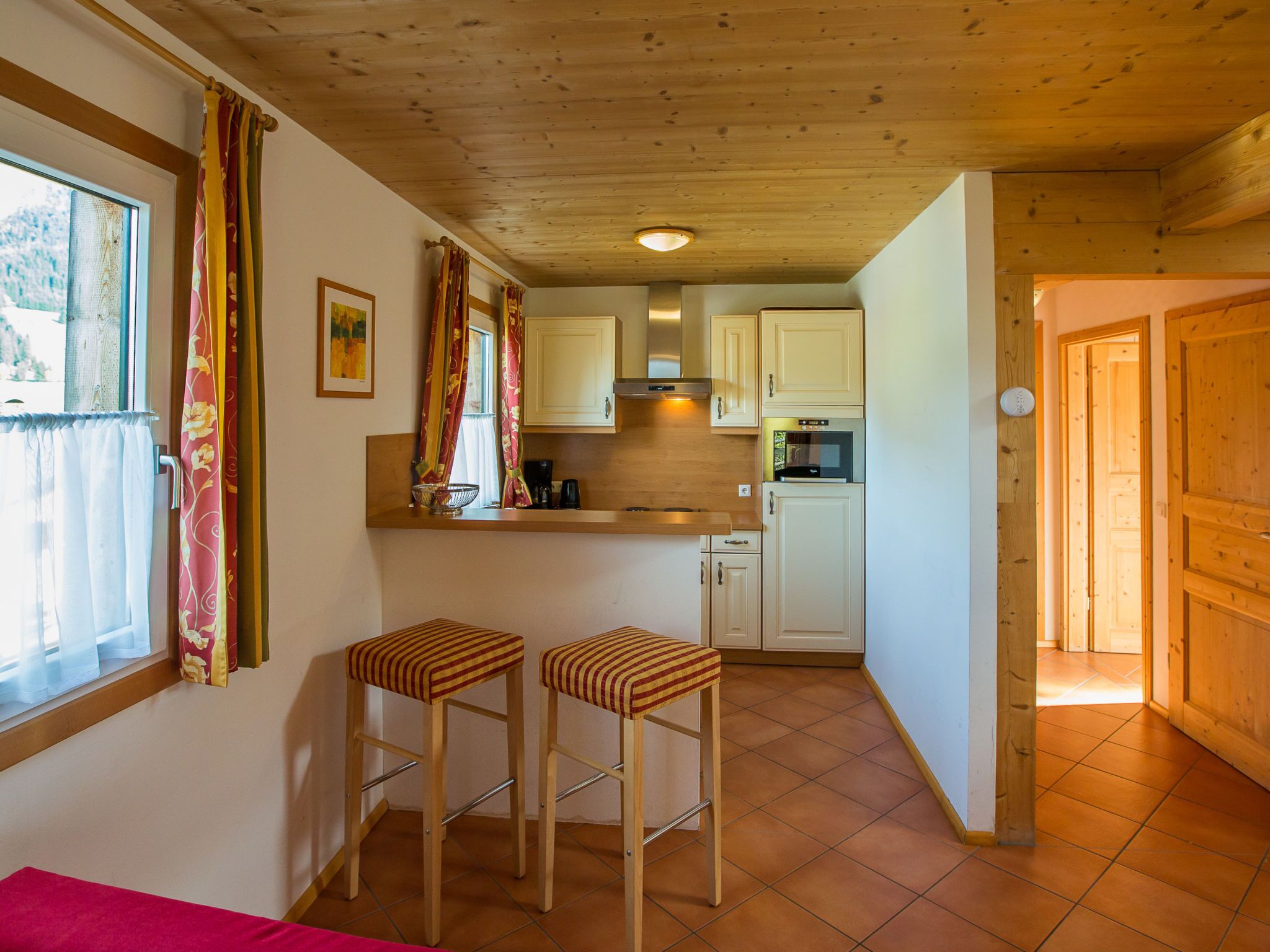 Cocina compacta con armarios de madera, horno integrado y cafetera. Hay dos taburetes a rayas cerca de la ventana.