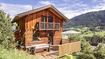 Eine Holzhütte mit Balkon, umgeben von viel Grün und einer Berglandschaft.