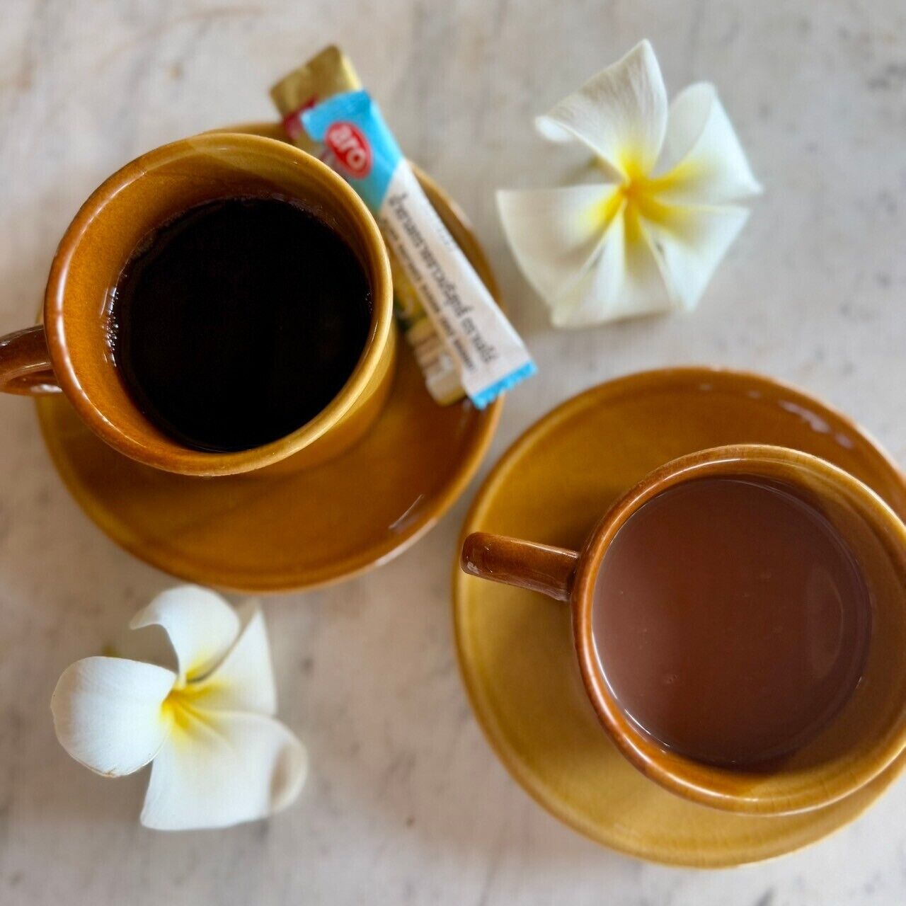 Two cups of coffee on a table with a packet of sugar and a flower.