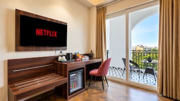 A hotel room with a wooden desk, a red chair, a minibar, a television with 'NETFLIX' on the screen, and a balcony with a view.