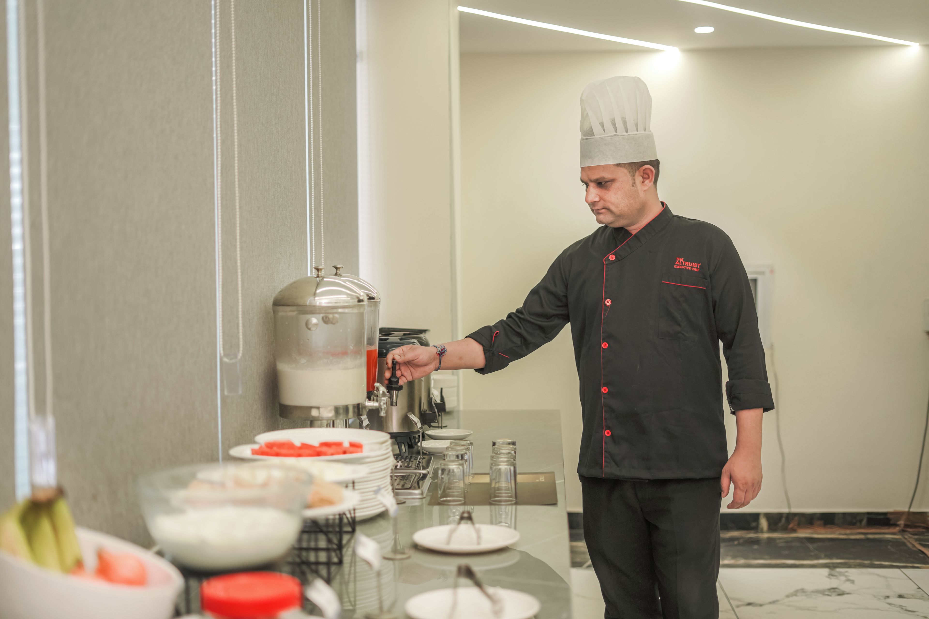 Un chef con uniforme negro y detalles rojos, de pie en un área de cocina con un dispensador de bebidas y varios platos sobre el mostrador.