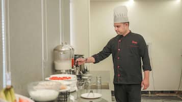 Un chef con uniforme negro y detalles rojos, de pie en un área de cocina con un dispensador de bebidas y varios platos sobre el mostrador.