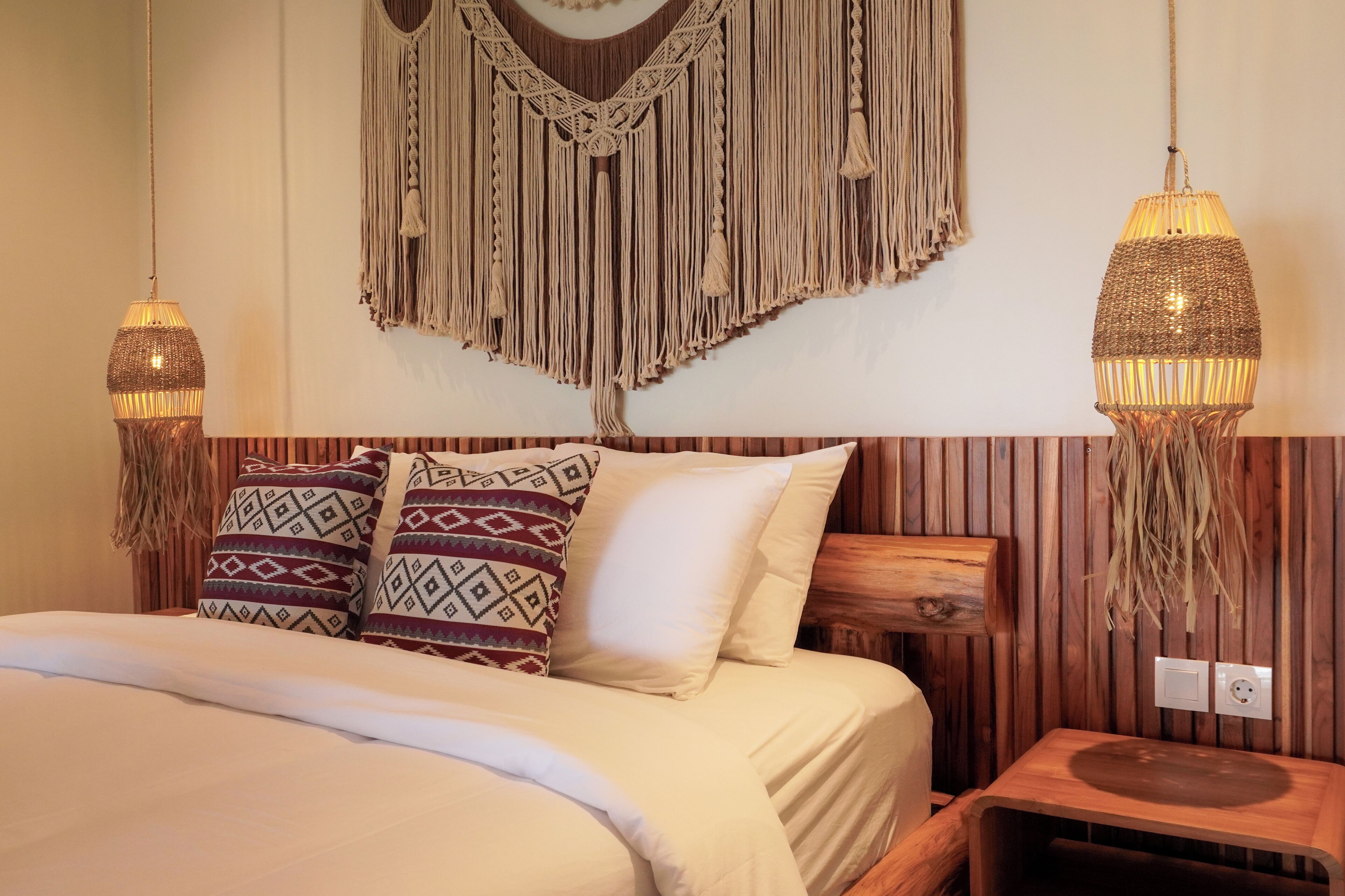 Un lit avec des coussins à motifs, une décoration murale en macramé et deux lampes suspendues.