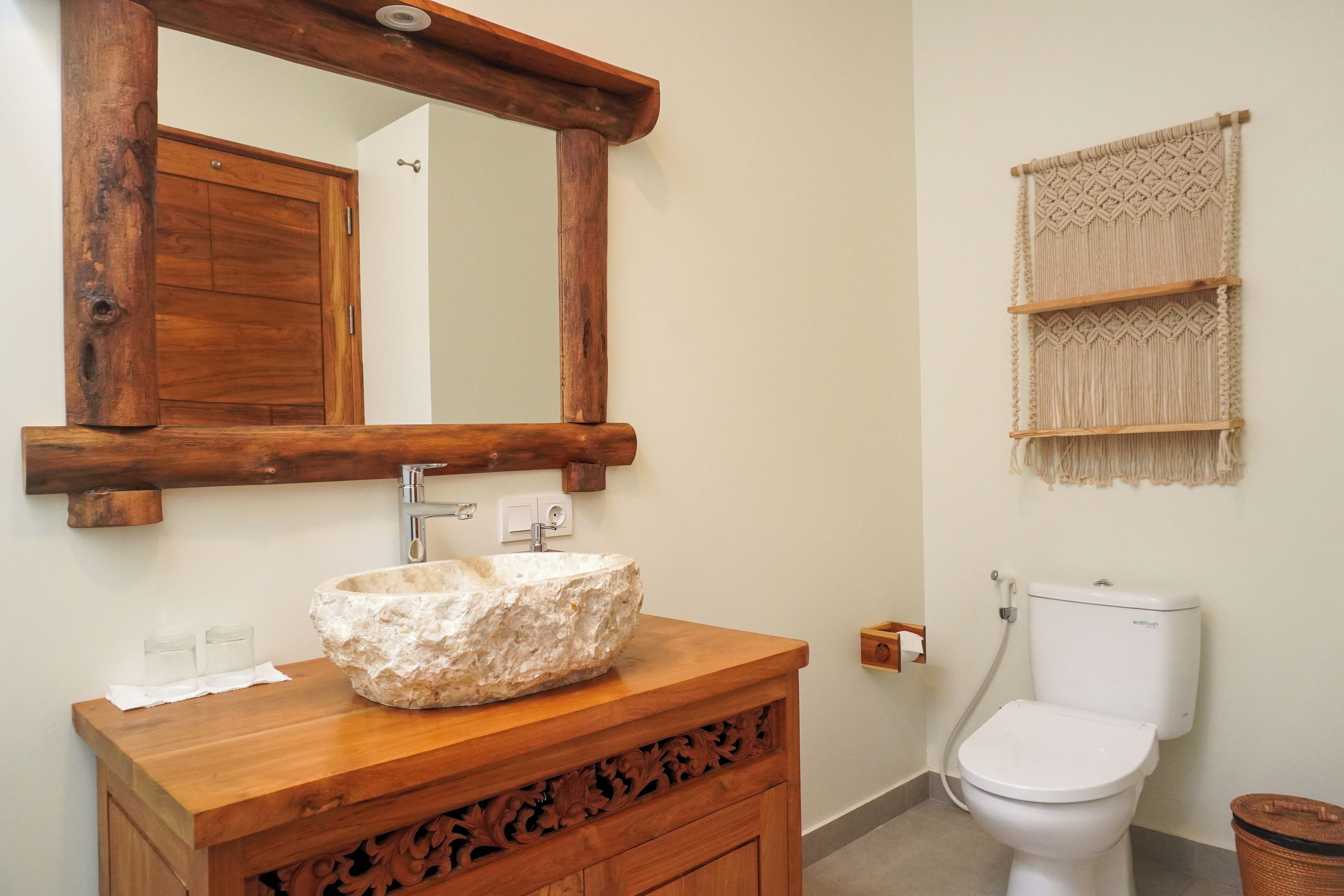 A bathroom with a wooden vanity, a stone sink, a wooden-framed mirror, a toilet, and a macramé wall hanging.