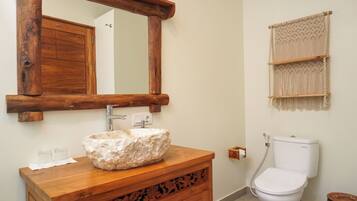 A bathroom with a wooden vanity, a stone sink, a wooden-framed mirror, a toilet, and a macramé wall hanging.