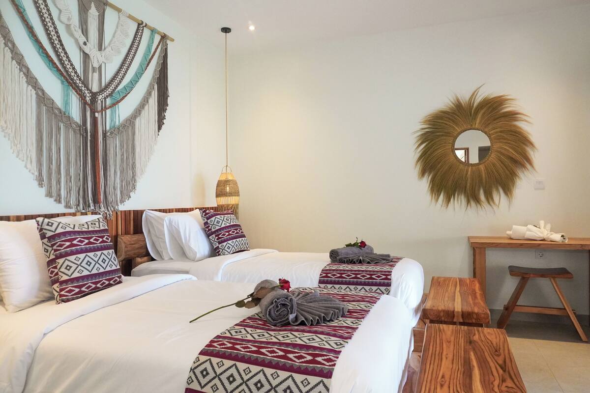 Ein Hotelzimmer mit zwei Betten, einem Holztisch, einem Spiegel und einer Wanddekoration aus Makramee.