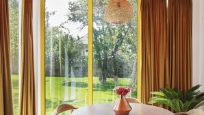 A dining area with a round table, four chairs, a vase with flowers, and a view of a garden through large windows. - Major's House Hotel (Step'anavan)