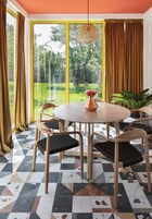 Un comedor con una mesa redonda, cuatro sillas, un jarrón con flores y vistas a un jardín a través de amplias ventanas.
