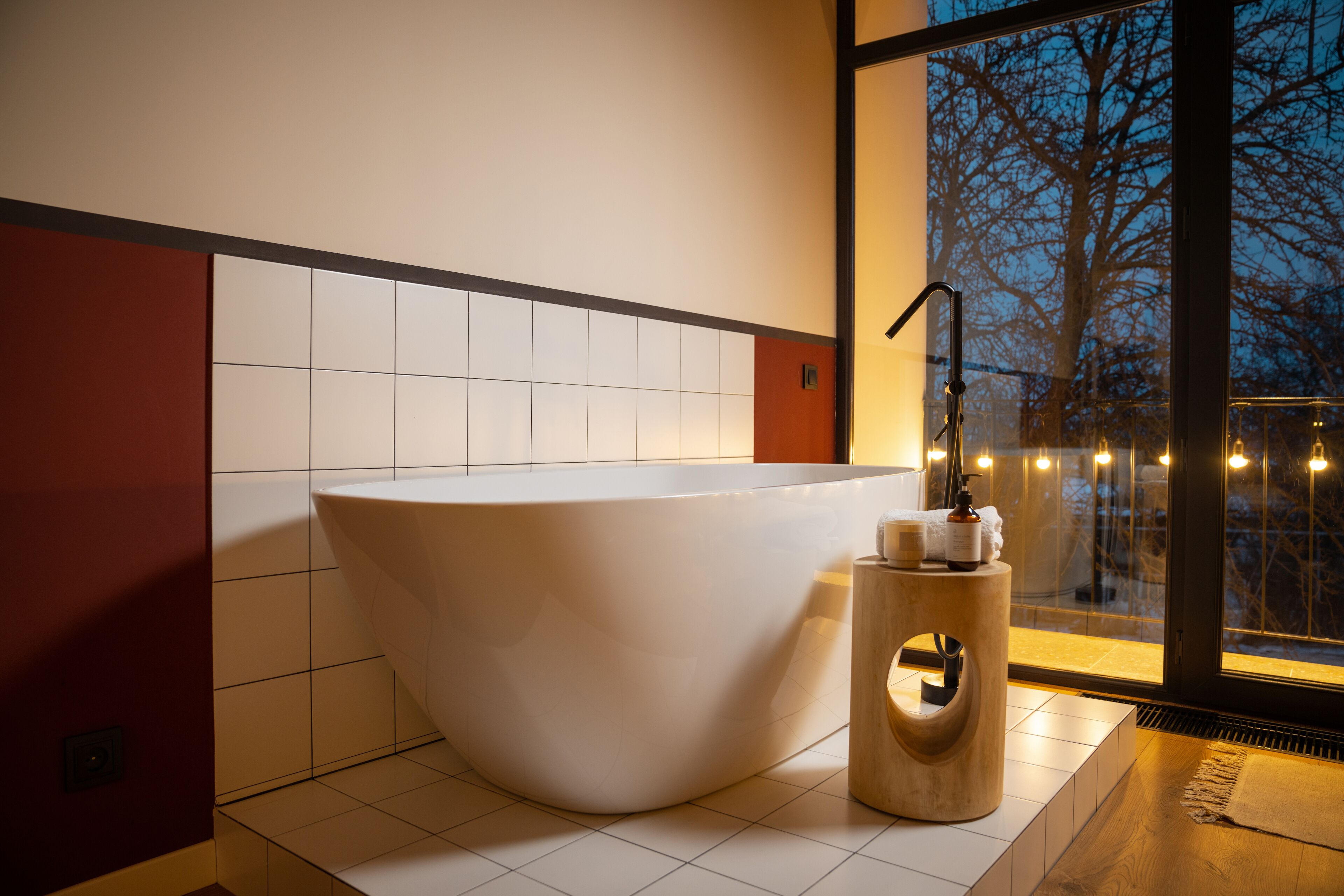 A modern bathroom with a freestanding bathtub, a wooden side table, and a view of trees through a large window.