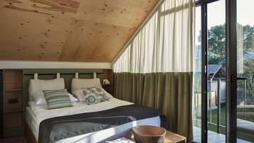 A bedroom with a sloped ceiling, a large bed with pillows and a blanket, a bedside table with towels, and a view through a glass door to the outdoors.
