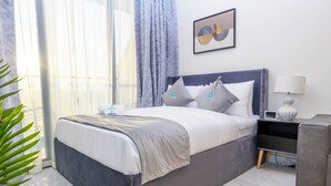 A modern hotel room with a large bed, a bedside table with a lamp, and a view of the outdoors through sheer curtains.