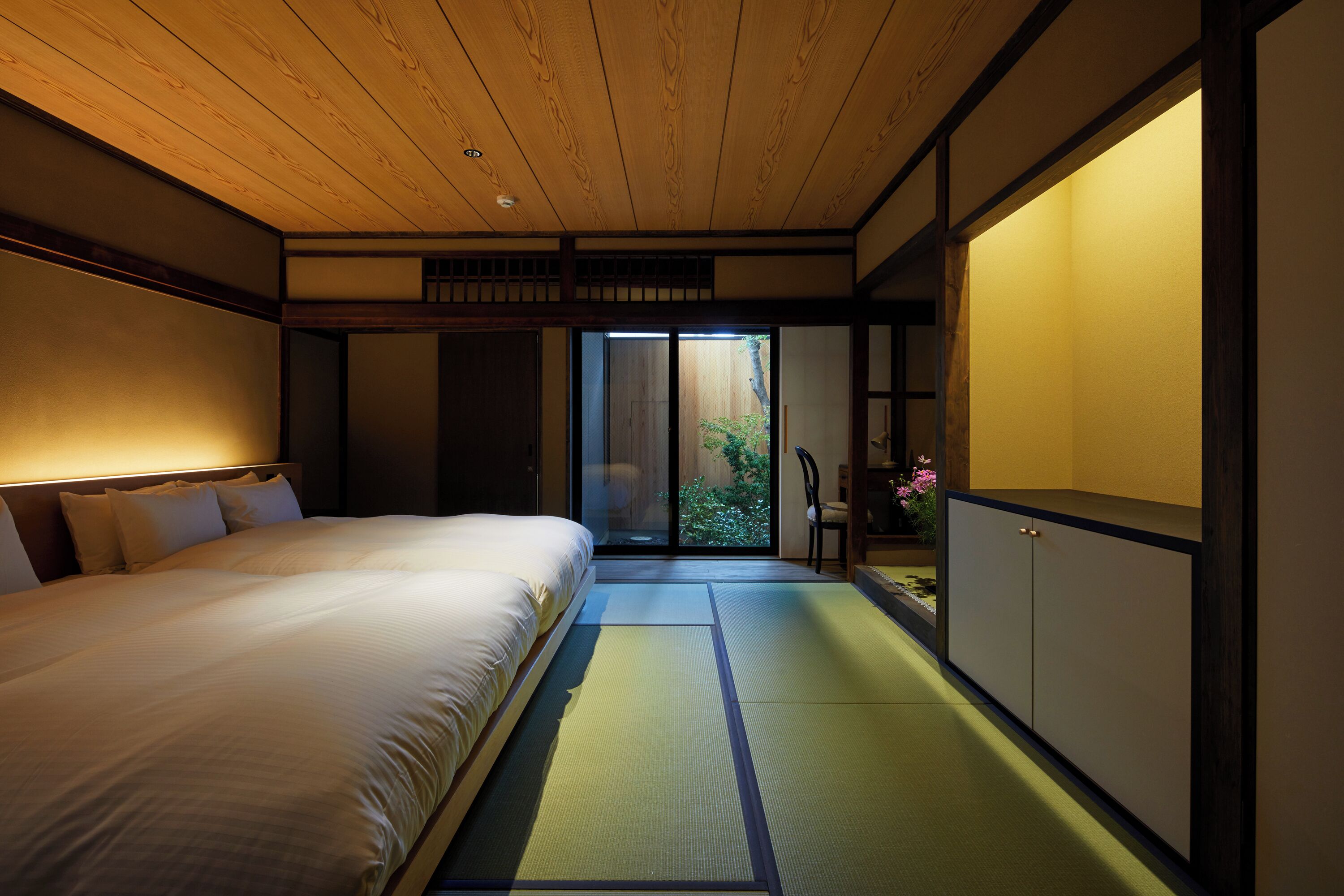 A traditional Japanese-style room with tatami flooring, a low wooden bed, sliding doors, and a view of greenery outside.