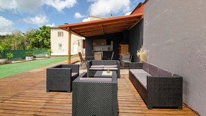 An outdoor patio with wicker furniture, a wooden deck, and a covered area with a kitchen in the background. - Club Villamar - Kaixo is a detached Spanish villa with a private pool, garden, BBQ, spacious livi... (Calonge)