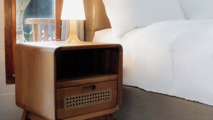 A wooden bedside table with a lamp, a drawer, and a view of a bed with white bedding. - DMY Guest House Nusa Penida (Penida Island)