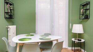 Un comedor con una mesa blanca, sillas verdes y un radiador blanco. Hay dos estanterías negras con objetos decorativos en las paredes.