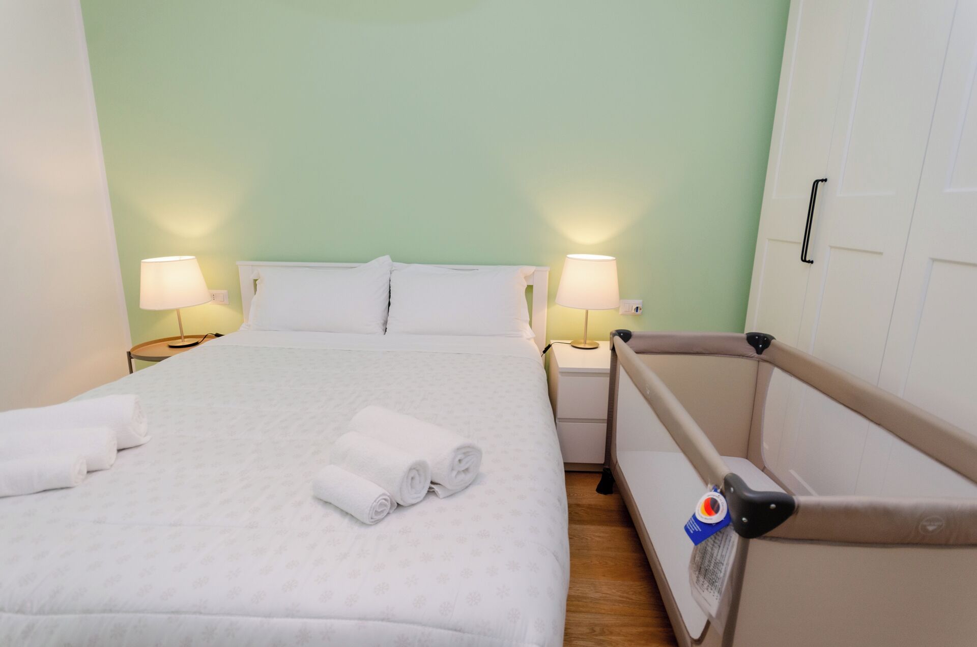 Een hotelkamer met een bed, twee bedlampen, een nachtkastje en een babybed met een veiligheidshek.