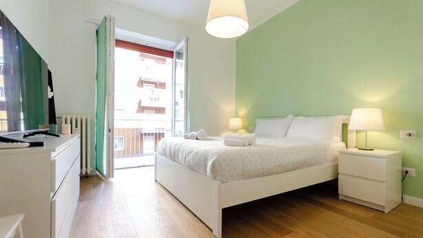 Un dormitorio con cama, mesitas de noche, cómoda y ventana con vistas a edificios.