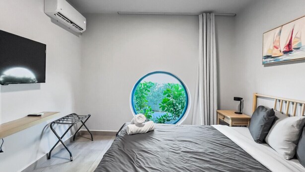 A modern hotel room with a large bed, a round window with a view of greenery, a flat-screen TV mounted on the wall, and a small bedside table with a lamp.