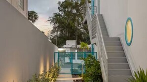 A narrow pathway leading to a staircase with a blue circular logo, surrounded by greenery and a white building with a grey roof.