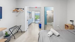 Ein modernes Hotelzimmer mit Bett, kleinem Schreibtisch, Pflanze und Blick auf das Badezimmer und die Küche.