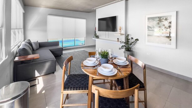 Una sala de estar moderna con comedor, un sofá gris, un televisor de pantalla plana y vista al océano.